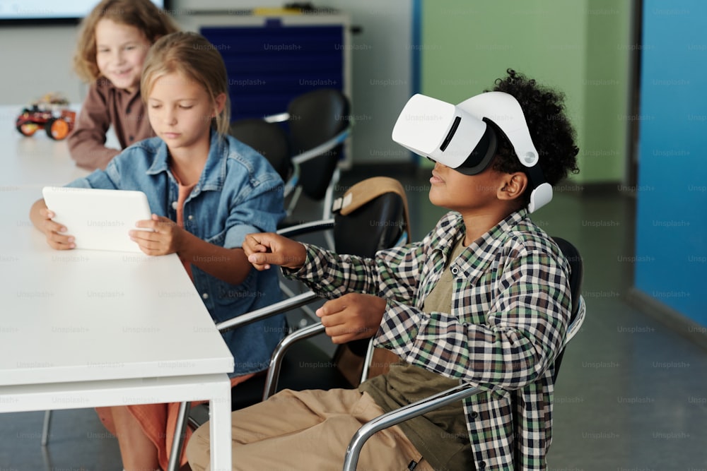 Vue latérale d’un écolier afro-américain mignon dans un casque VR contrôlant un robot virtuel pendant que son camarade de classe regarde l’action sur l’écran de la tablette