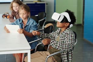 Vista lateral do estudante afro-americano bonito no fone de ouvido vr controlando o robô virtual enquanto seu colega de classe assistindo ação na tela do tablet