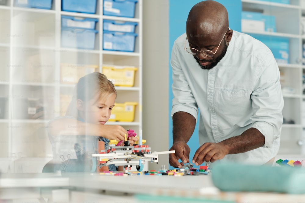 Jovem negro de camisa casual branca ajudando aluna a construir robô enquanto está ao lado dela e se curvando sobre a mesa