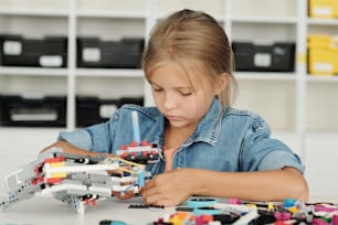 Cute blond schoolgirl in denim shirt connecting parts of electronic robot while sitting by desk at lesson and doing what teacher says