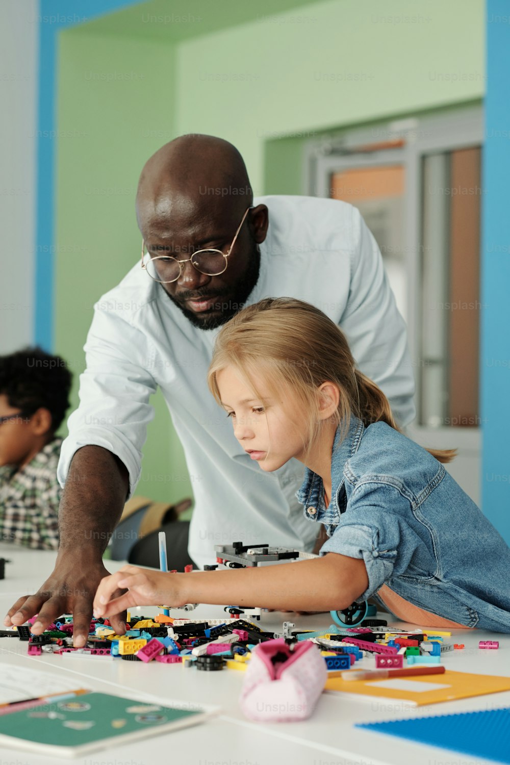 Cute schoolgirl choosing details for construction of robot with help of teacher standing next to her and bending over workplace