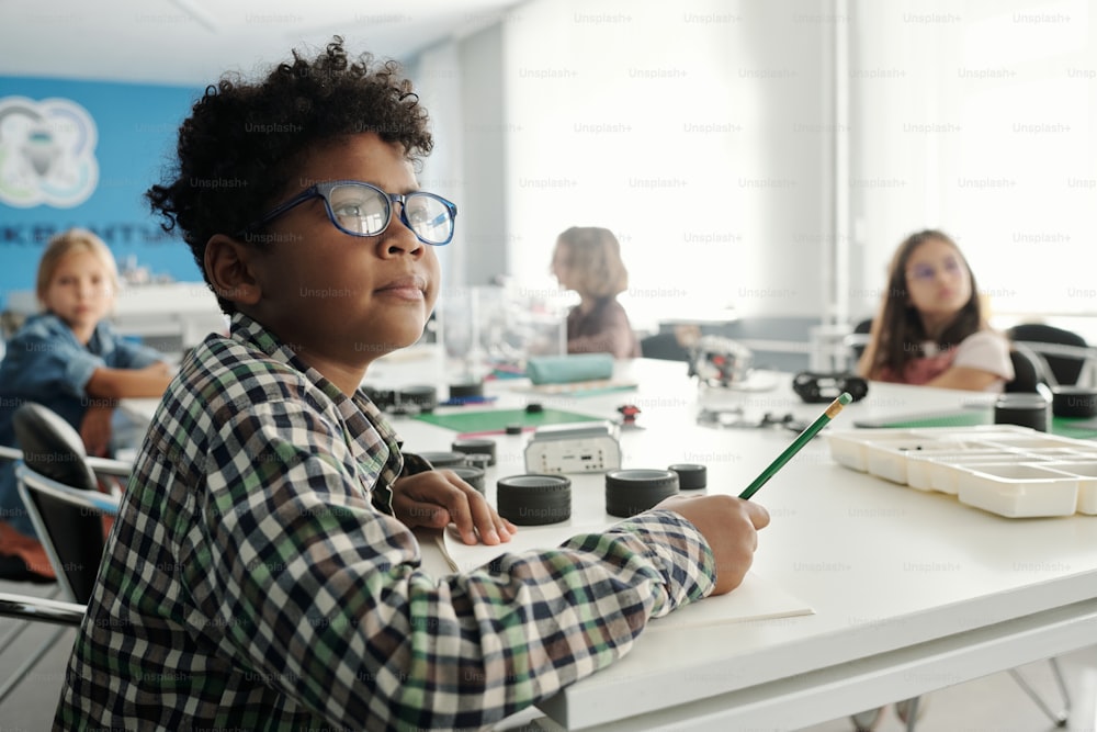 Un écolier intelligent portant des lunettes et une chemise à carreaux prend des notes dans un cahier de notes tout en étant assis à table contre ses camarades de classe