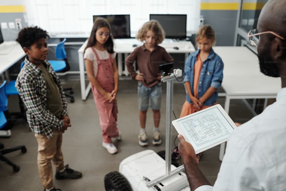 African American teacher with tablet standing in front of schoolkids and robot crawler while making presentation of new toy at lesson