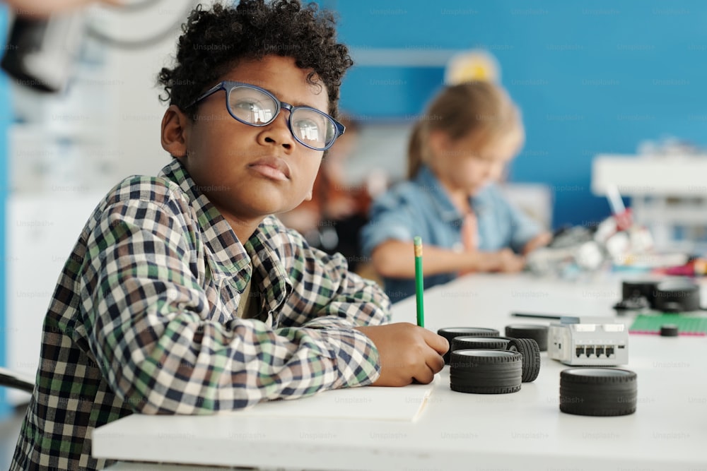 안경을 쓴 진지한 젊은이가 로봇 수업에서 선생님을 바라보고 로봇의 부품으로 책상 옆에 앉아 메모를 하고 있다