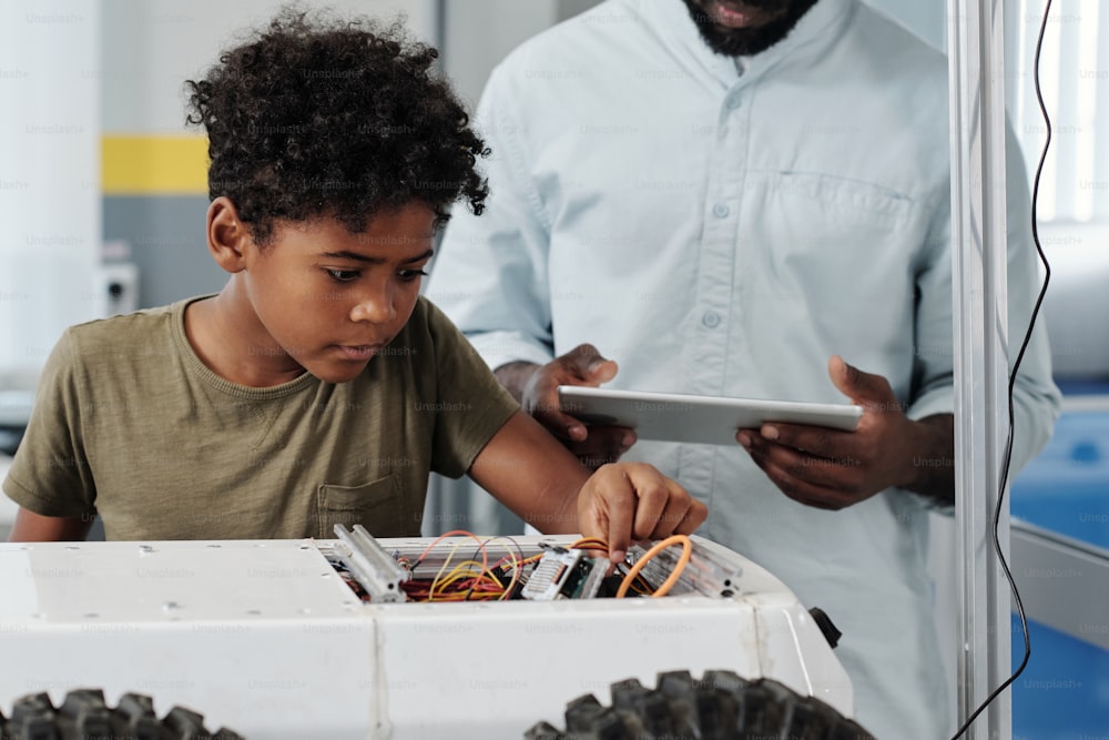 Lindo colegial afroamericano conectando cables de robot de juguete mientras sigue las instrucciones de su profesor de robótica con tableta