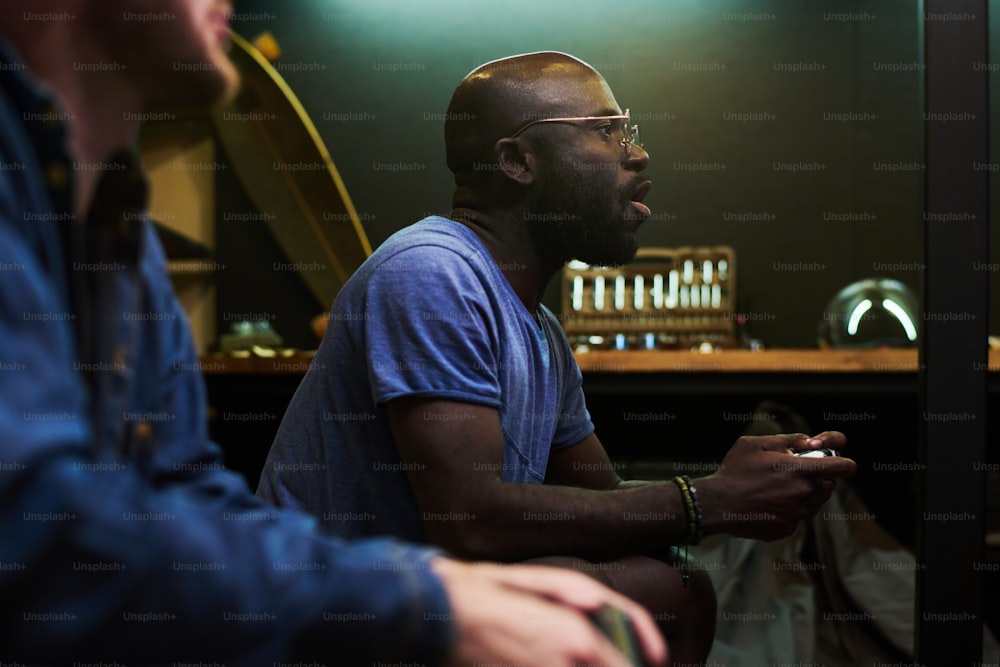 Junger begeisterter schwarzer Mann mit Gamepad, der während des Videospiels auf der Couch vor dem Fernsehbildschirm sitzt und die Tasten am Joystick drückt