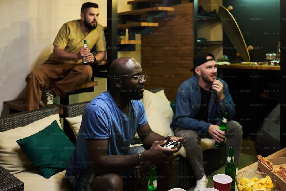 Two young men with beer looking at tv screen and watching video game while black guy with joystick sitting near by and pressing buttons