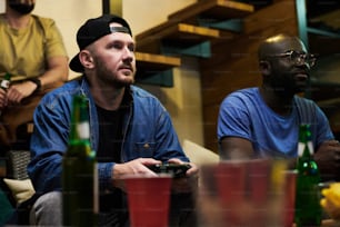 Young serious man and his buddy with gamepads concentrating on leisure game while looking at screen of tv set