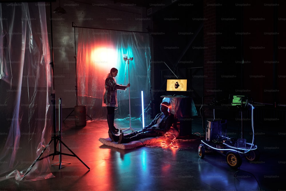 Jeune femme réparant un tuyau compte-gouttes sur le bras d’un homme cyberpunk alors qu’elle est assise par terre dans une pièce sombre avec des néons