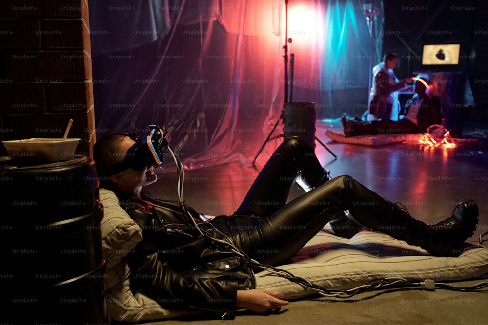 Giovane donna in costume di pelle sdraiata sul materasso in occhiali di realtà virtuale, persone che praticano l'intelletto artificiale nella stanza buia sullo sfondo