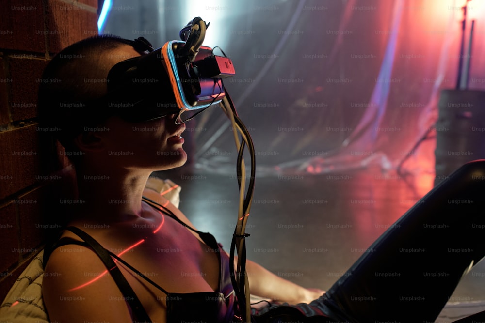 Young cyberpunk woman in virtual reality goggles playing online game in dark room