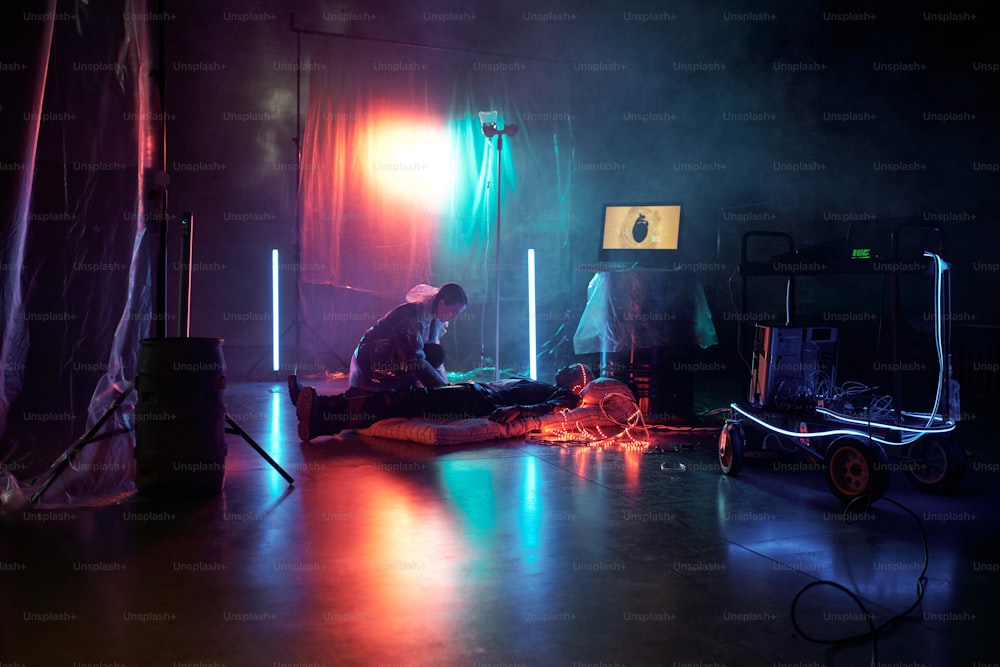 Mujer joven conecta al hombre con la realidad virtual mientras está acostado en un colchón en una habitación oscura