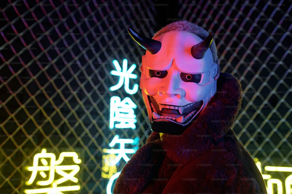 Close-up of cyberpunk guy in devil mask and fur coat looking at camera standing against the bars and hieroglyphs meaning nirvana and western paradise