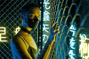 Portrait of cyberpunk woman with black paint on her face standing near the bars and looking at camera with hieroglyphs meaning nirvana and western paradise