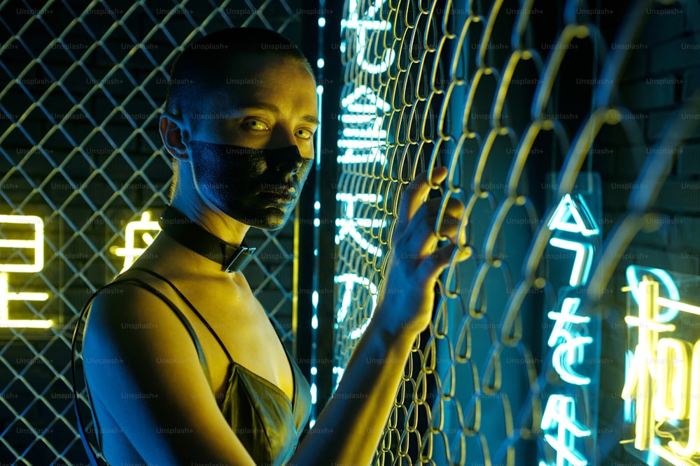 Portrait of cyberpunk woman with black paint on her face standing near the bars and looking at camera with hieroglyphs meaning nirvana and western paradise