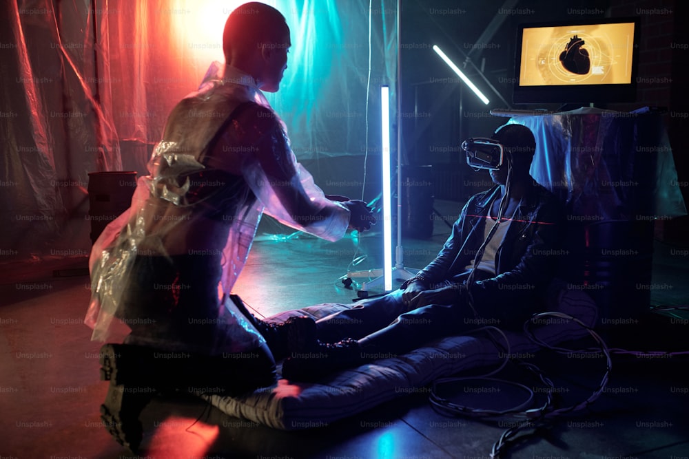 Femme en manteau de protection contrôlant la réalité virtuelle sur l’écran de l’ordinateur tandis que l’homme est assis dans des lunettes VR et joue à un jeu en ligne
