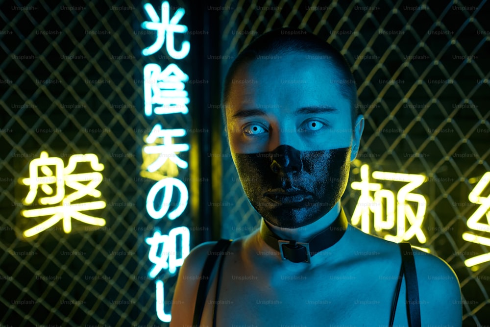 Young cyberpunk female with her face half painted in black standing in front of camera against bars and illuminated hieroglyphs