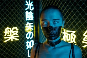 Young cyberpunk female with her face half painted in black standing in front of camera against bars and illuminated hieroglyphs