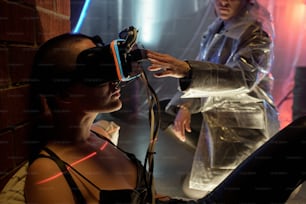 Hand of young woman touching virtual reality headset on head of cyberpunk girl in black leather pants and tanktop sitting by brick wall
