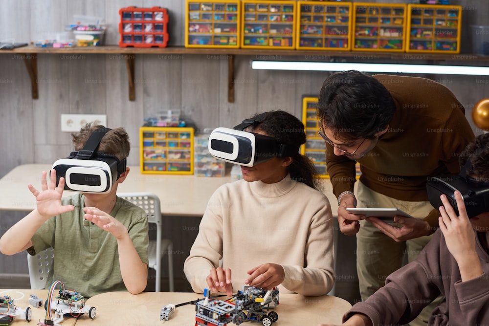 Group of children sitting at the table and projecting robots in vr glasses with teacher assisting them during robotics lesson
