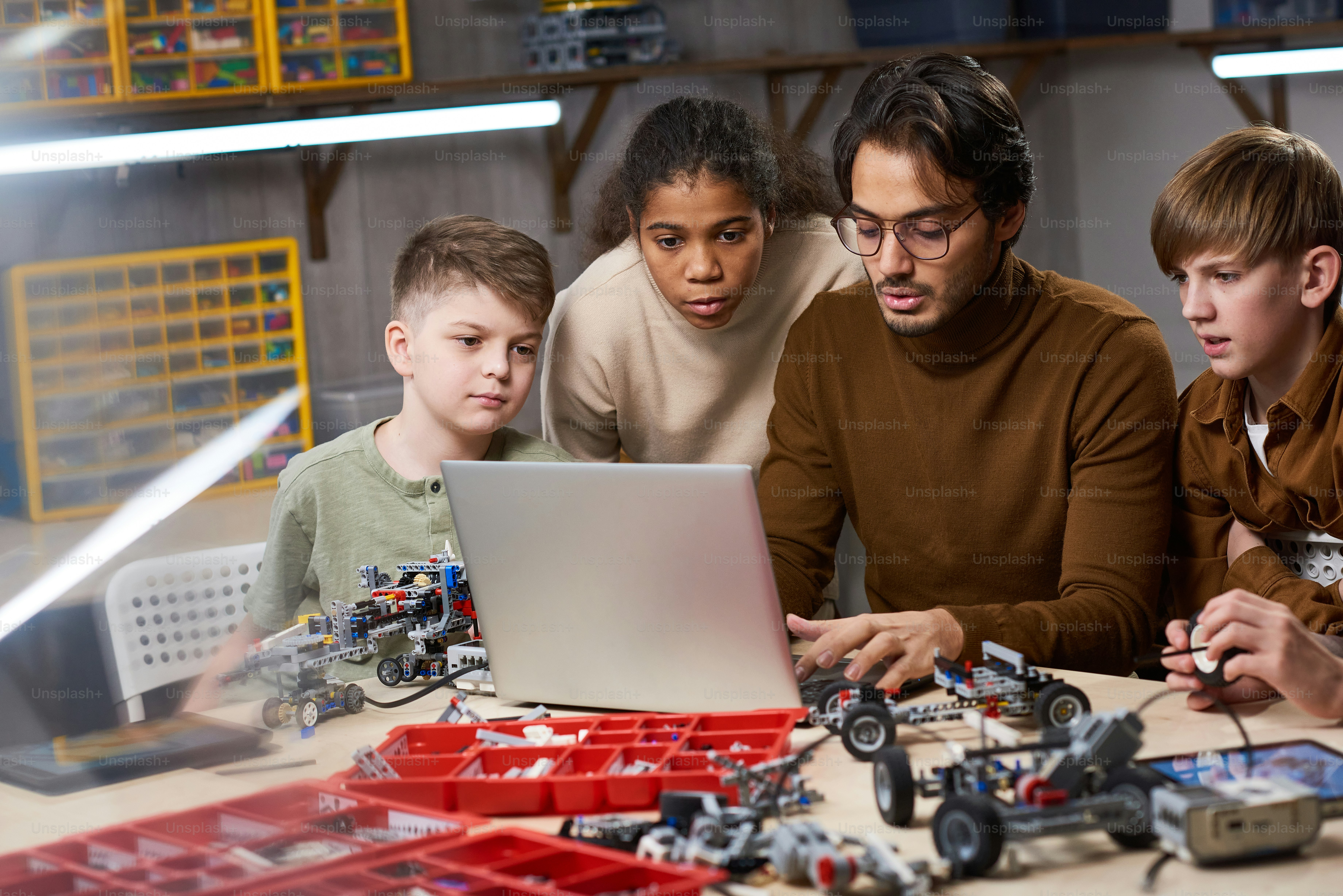 Sekelompok anak-anak melihat monitor laptop saat guru mengetik di laptop, dia menunjukkan bagaimana memproyeksikan robot