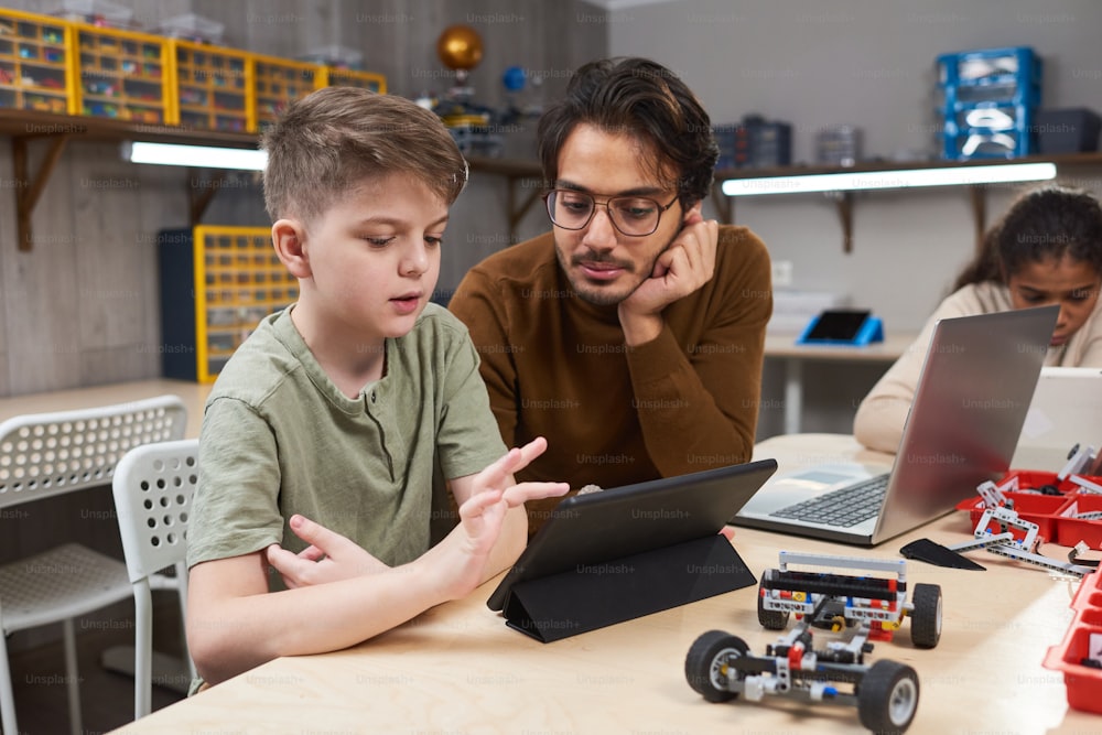 로봇 수업에서 컴퓨터로 로봇을 제어하는 교사와 함께 테이블에 앉아 디지털 태블릿을 사용하는 소년