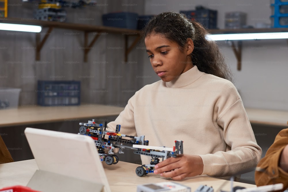 African girl sitting at the table in front of the tablet pc and making robot from constructor at science school