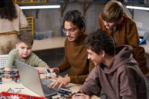 Jovem de óculos sentado à mesa e digitando no laptop, ele ensina os alunos a se conectarem on-line com robôs
