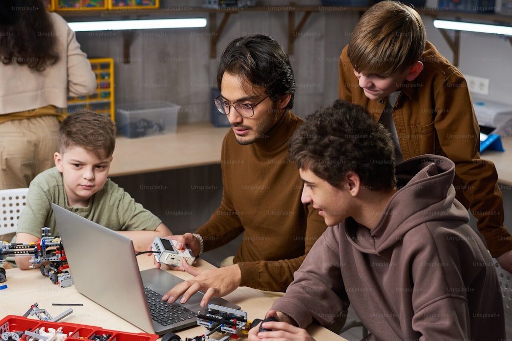 Young man in eyeglasses sitting at the table and typing on laptop, he teaching students to connecting online with robots