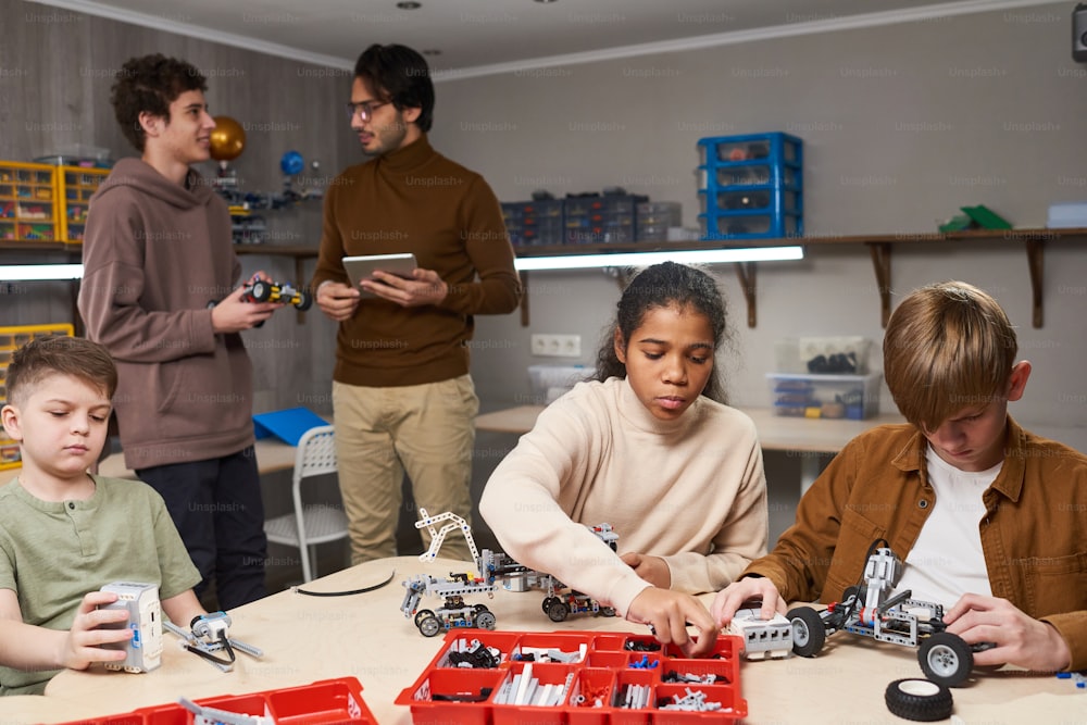 Grupo de adolescentes sentados à mesa e construindo robôs do construtor com o professor em pé no fundo na aula de robótica