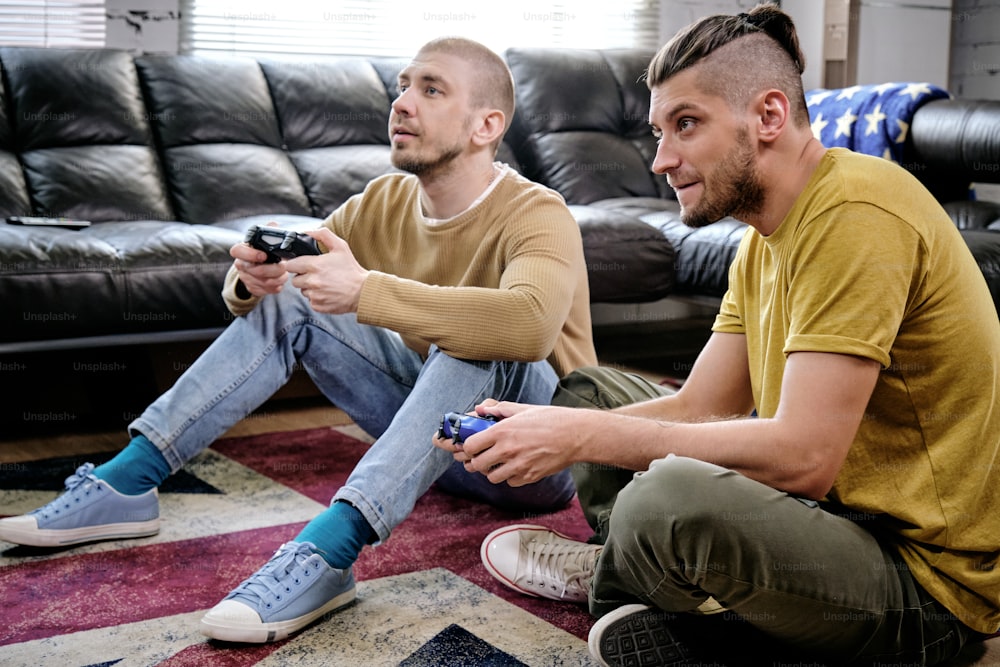 Two guys with joysticks playing video game on the floor in living-room of apartment they are sharing