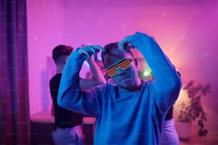 Young serious man in casualwear and disco eyeglasses looking at you while standing in front of his friends dancing together at home party