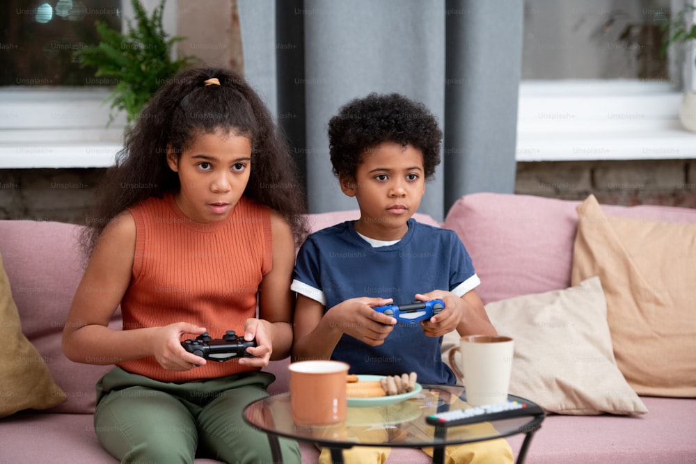 Cute boy and girl of African ethnicity sitting on couch against window in living-room and playing video game in front of tv set together