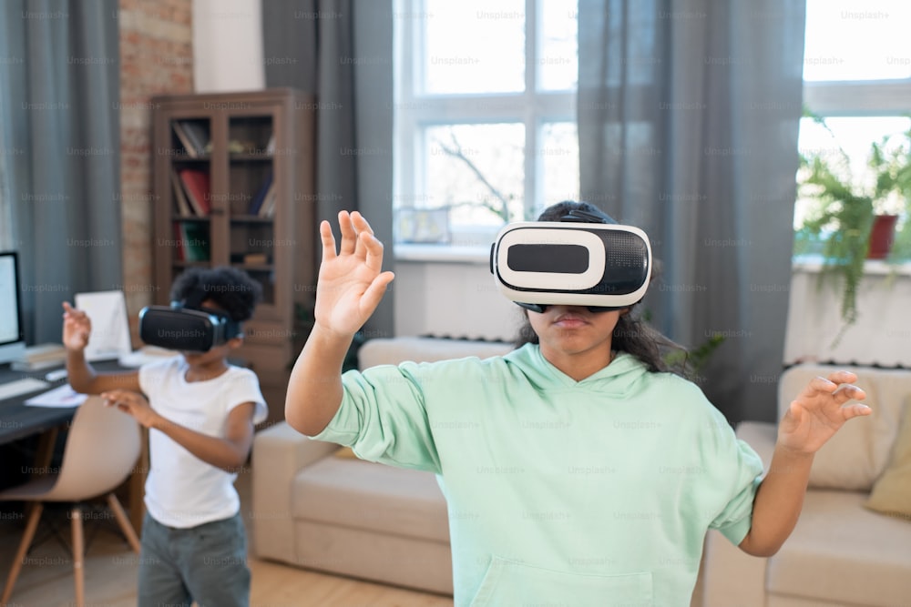Adorable mixed-race siblings in vr headsets watching online video on large virtual display while standing in front of camera in living-room