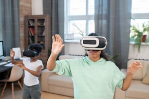 Adorable mixed-race siblings in vr headsets watching online video on large virtual display while standing in front of camera in living-room