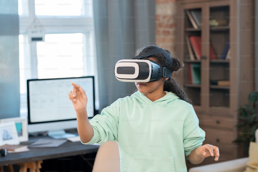 Adorable mixed-race girl in vr headset touching virtual display while watching online video in front of camera in home environment