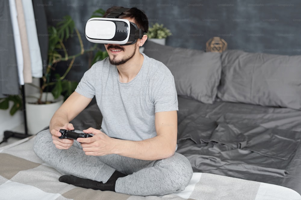 Jovem sorridente sentado com as pernas cruzadas na cama e jogando videogame usando joystick e fone de ouvido de realidade virtual
