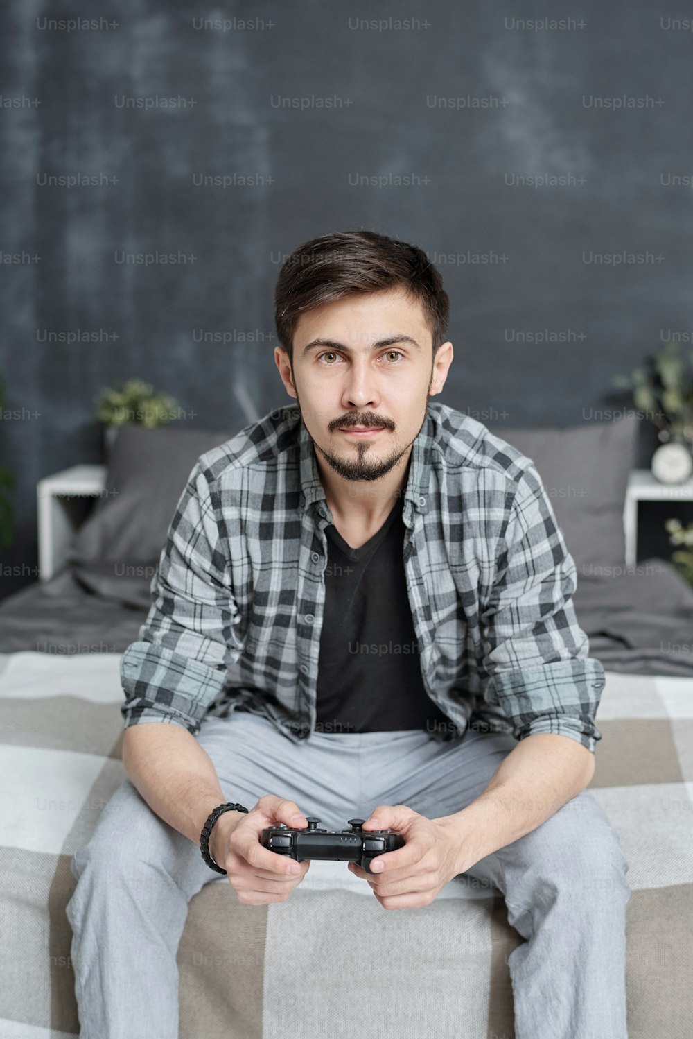 Retrato de un apuesto joven con camisa casual sentado en la cama y usando un joystick mientras juega un videojuego en casa