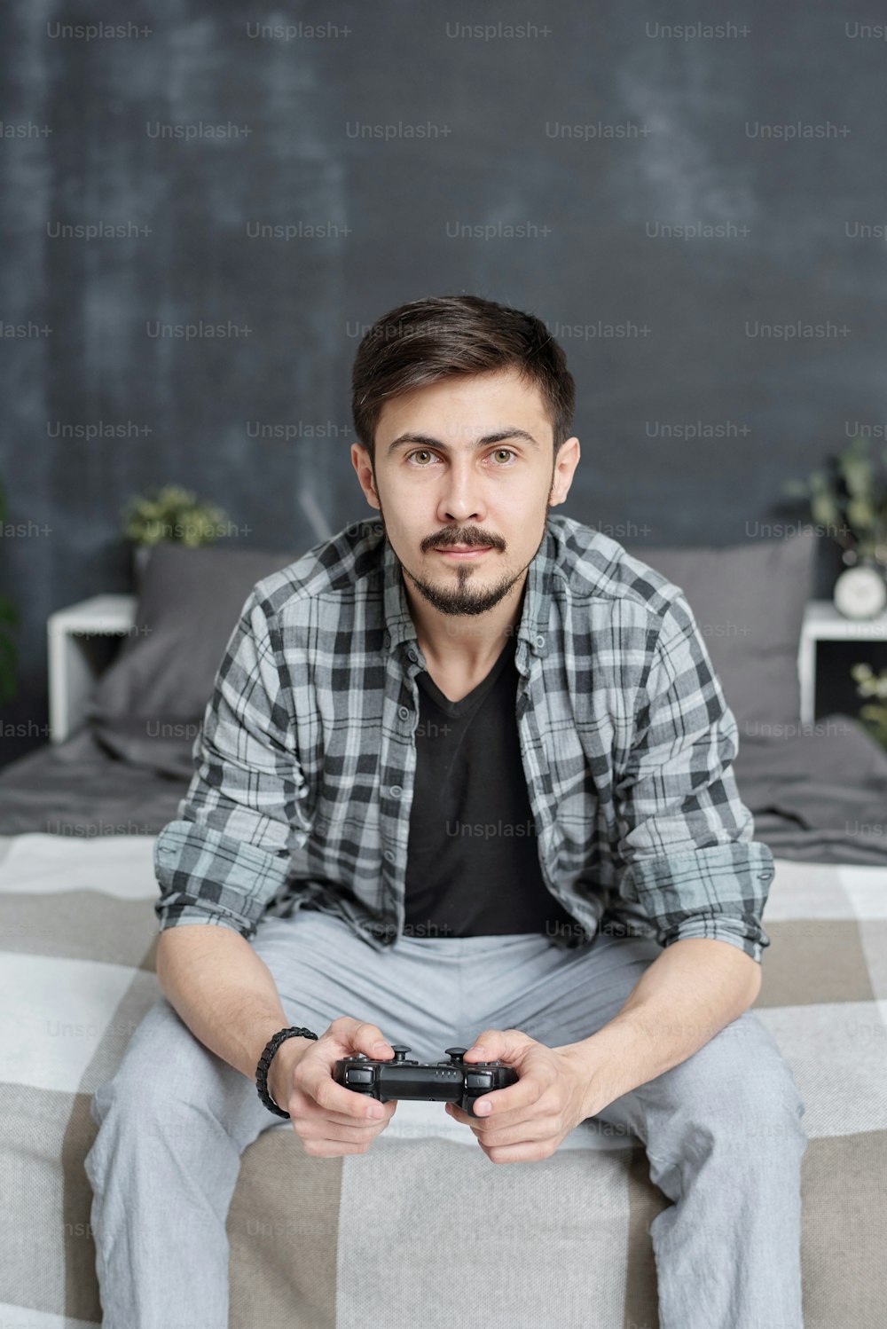 カジュアルなシャツを着たハンサムな若い男性がベッドに座り、自宅でビデオゲームをしながらジョイスティックを使う