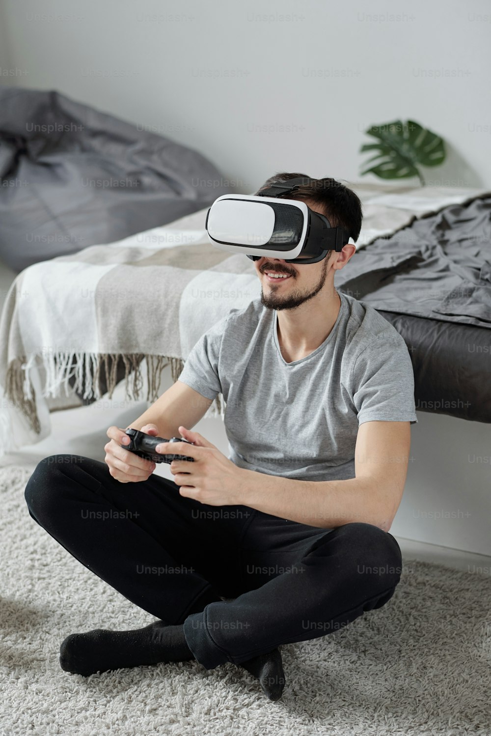 Jeune homme barbu joyeux assis les jambes croisées sur un tapis et jouant à un jeu vidéo en utilisant des lunettes de réalité virtuelle en quarantaine