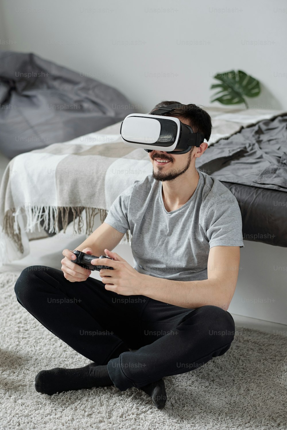 Cheerful young bearded man sitting with crossed legs on carpet and playing video game using virtual reality goggles at quarantine