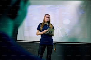 Young confident female developer of new network video game with digital tablet standing by large screen during presentation