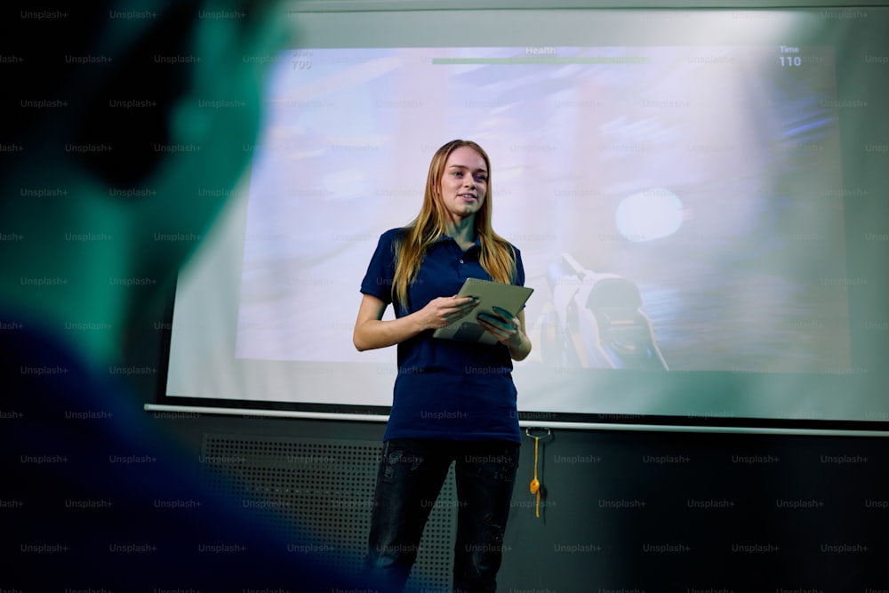 Joven desarrolladora de nuevos videojuegos en red con tableta digital de pie junto a una pantalla grande durante la presentación