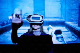 Young woman with vr goggle pushing virtual button while standing against large screen with projection of new e-sports video game