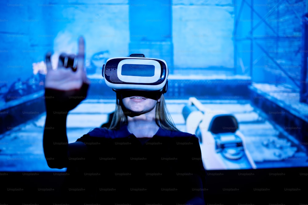Young woman with vr goggle pushing virtual button while standing against large screen with projection of new e-sports video game