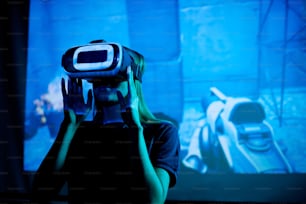 VR 헤드셋을 착용한 새로운 미래형 비디오 게임의 젊은 개발자가 프레젠테이션 중 가상 머신이 있는 대형 스크린에 서 있다