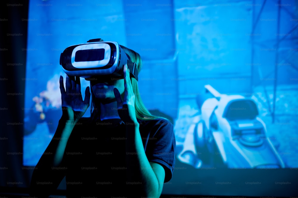 Junger Entwickler eines neuen futuristischen Videospiels mit VR-Headset, das während der Präsentation vor einem großen Bildschirm mit virtuellen Maschinen steht