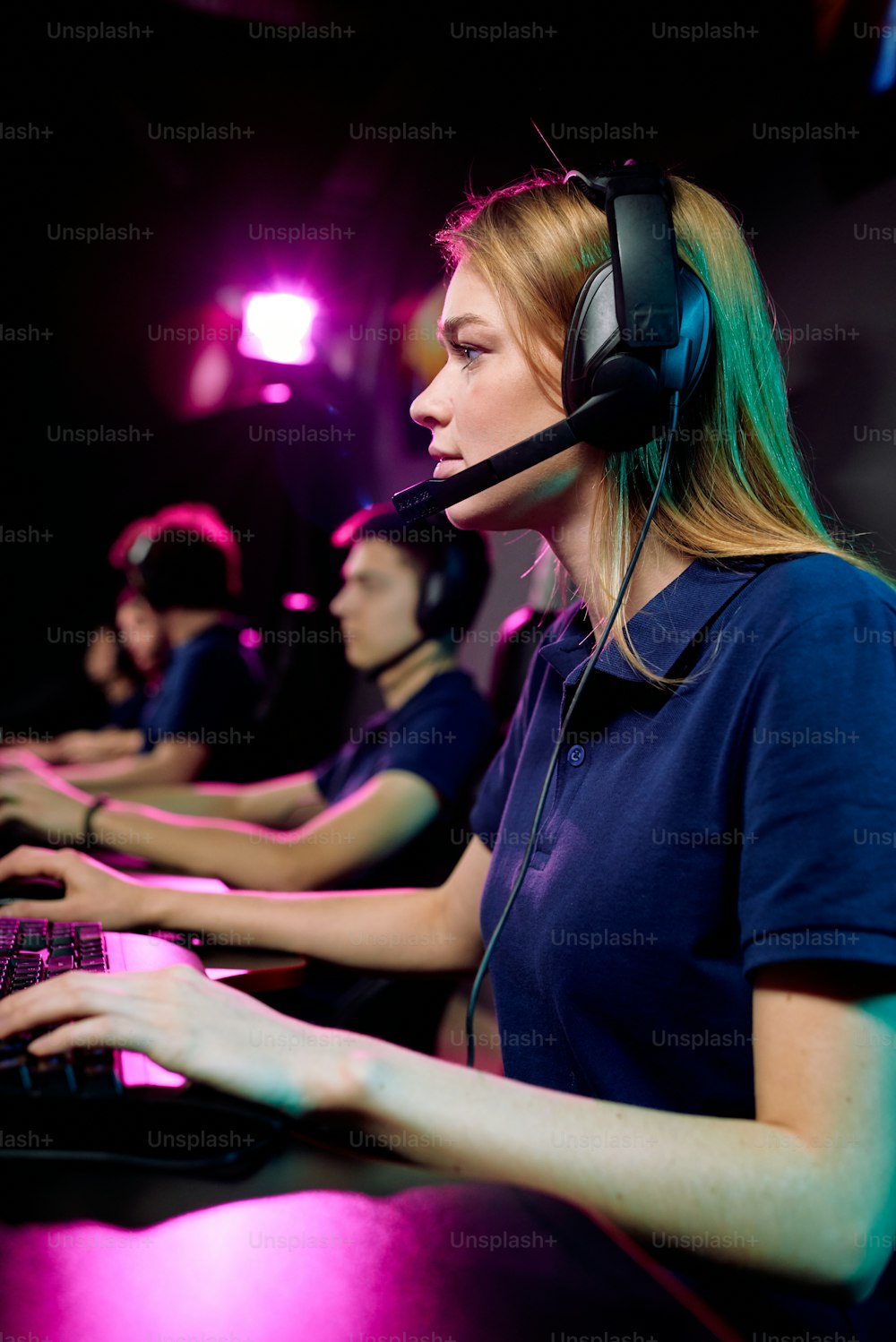 Focused young female computer gamer in hands-free headset with microphone using keyboard while playing online game in e-sports club