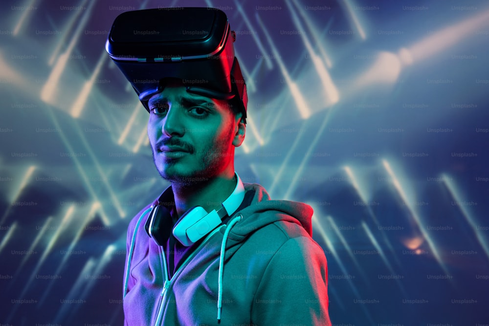 Portrait of serious young middle-eastern man with VR goggles on head standing against bright geometric background