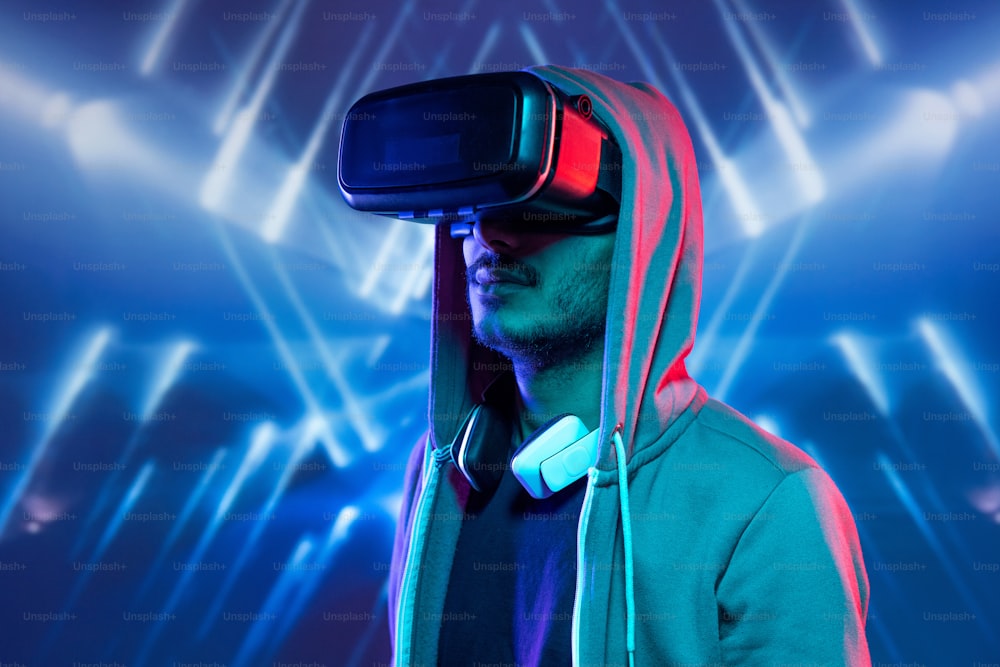 Hipster young man with stubble immersed in virtual reality wearing hoody sweatshirt watching 3D movie against blue luminous background